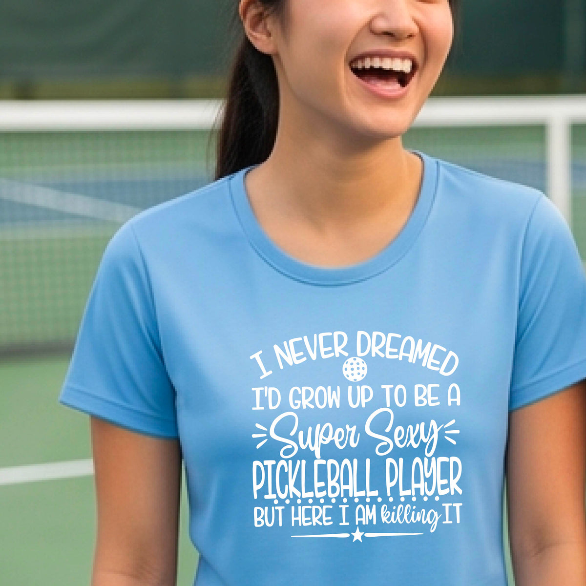 A woman on a pickleball court wearing a blue short-sleeve t-shirt with a white graphic of the text:  ‘I Never Dreamed I’d Grow Up To Be A Super Sexy Pickleball Player But Here I Am Killing It!’, standing on a pickleball court.
