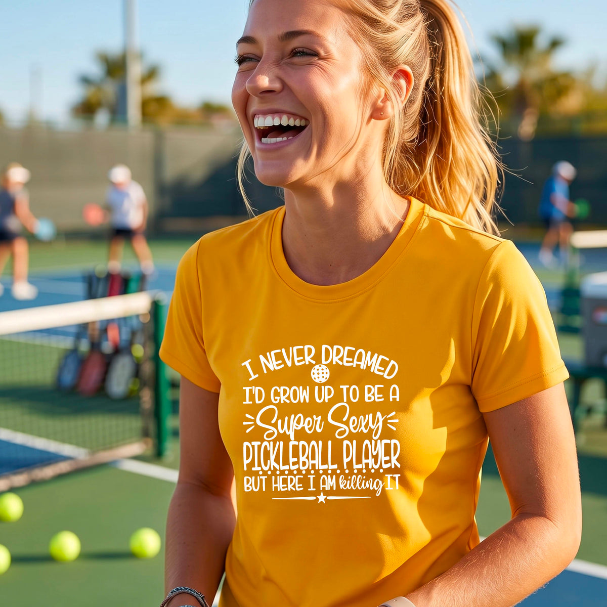 A woman on a pickleball court wearing a mustard yellow gold short-sleeve t-shirt with a white graphic of the text:  ‘I Never Dreamed I’d Grow Up To Be A Super Sexy Pickleball Player But Here I Am Killing It!’, standing on a pickleball court.