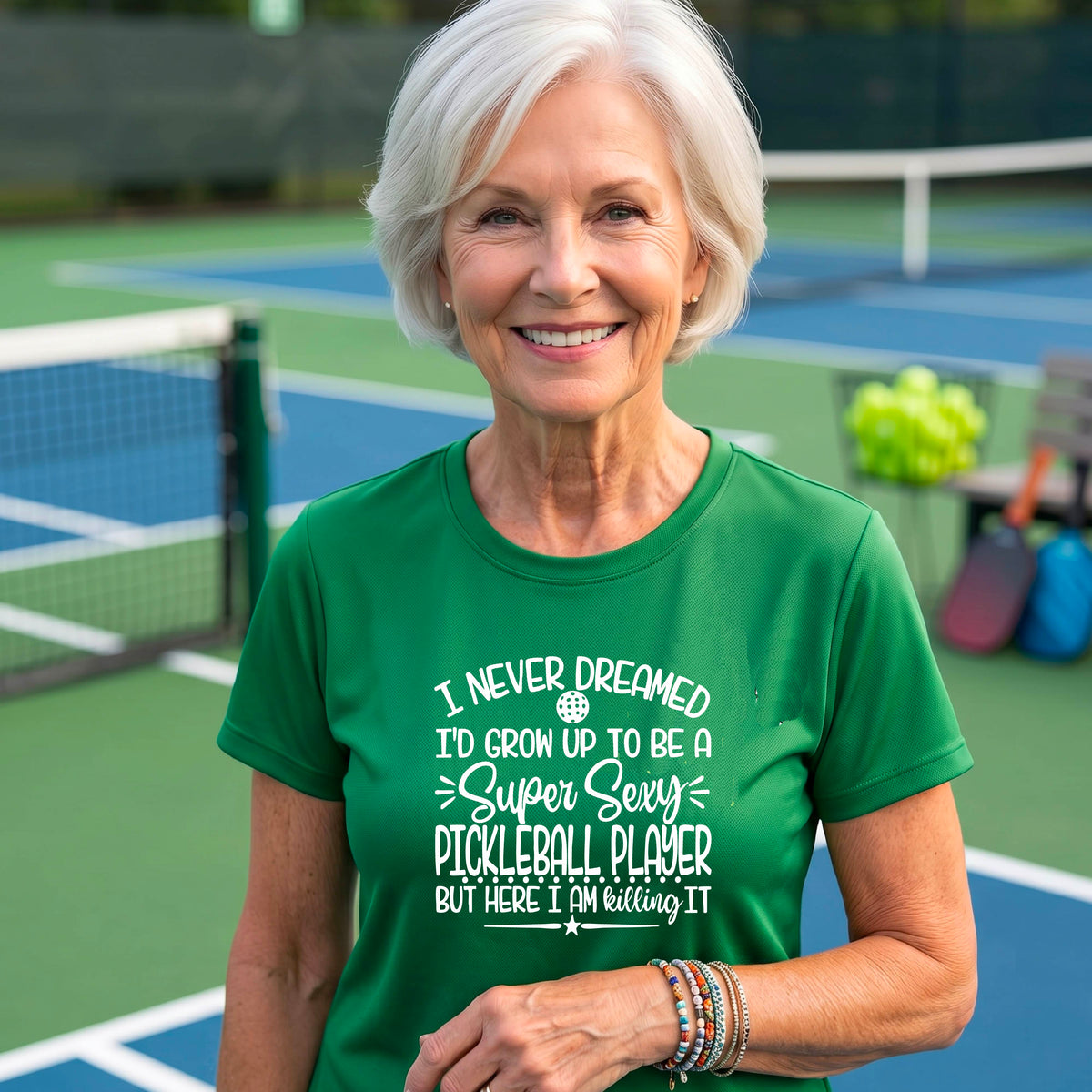 A woman on a pickleball court wearing a green short-sleeve t-shirt with a white graphic of the text:  ‘I Never Dreamed I’d Grow Up To Be A Super Sexy Pickleball Player But Here I Am Killing It!’, standing on a pickleball court.