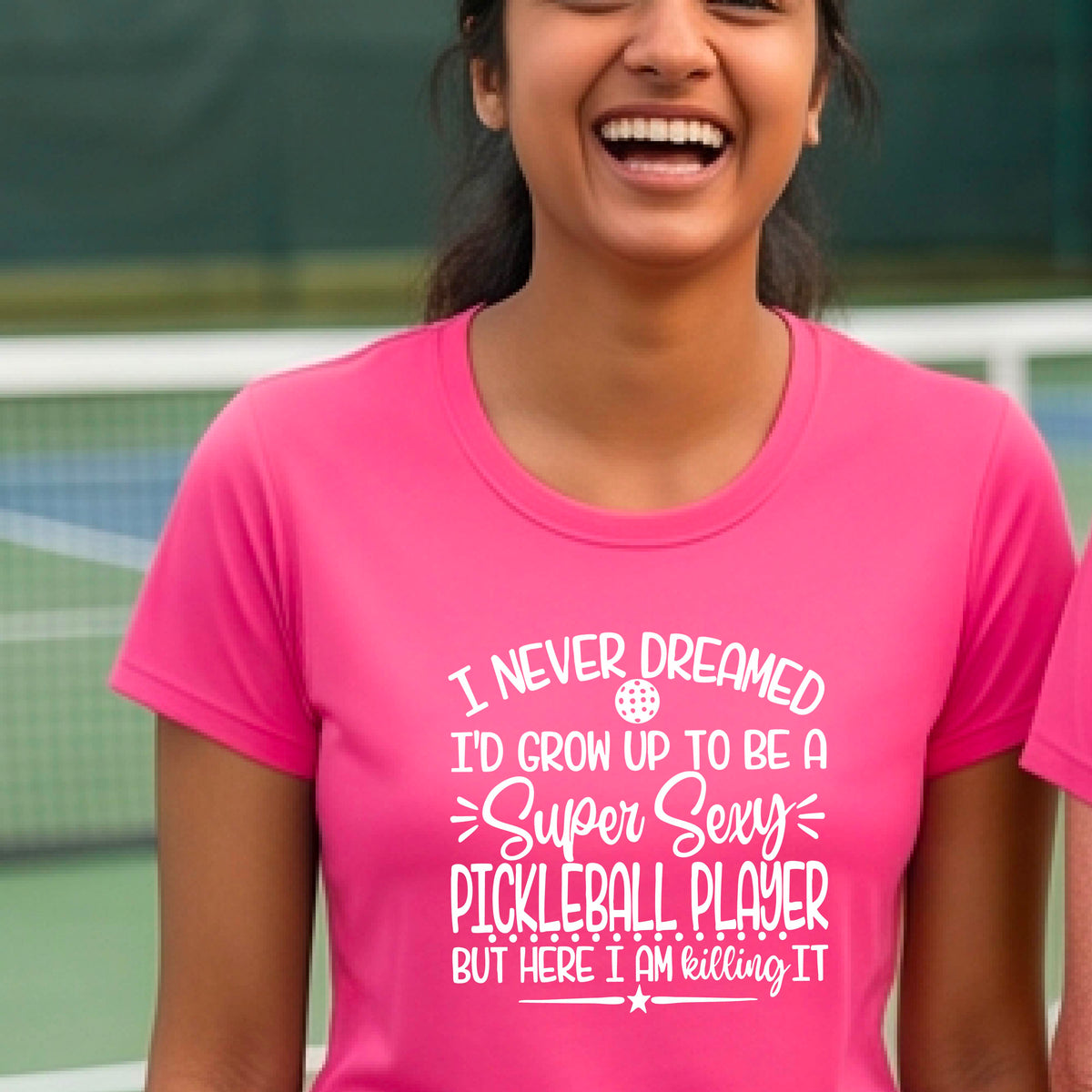 A woman on a pickleball court wearing a pink short-sleeve t-shirt with a white graphic of the text:  ‘I Never Dreamed I’d Grow Up To Be A Super Sexy Pickleball Player But Here I Am Killing It!’, standing on a pickleball court.