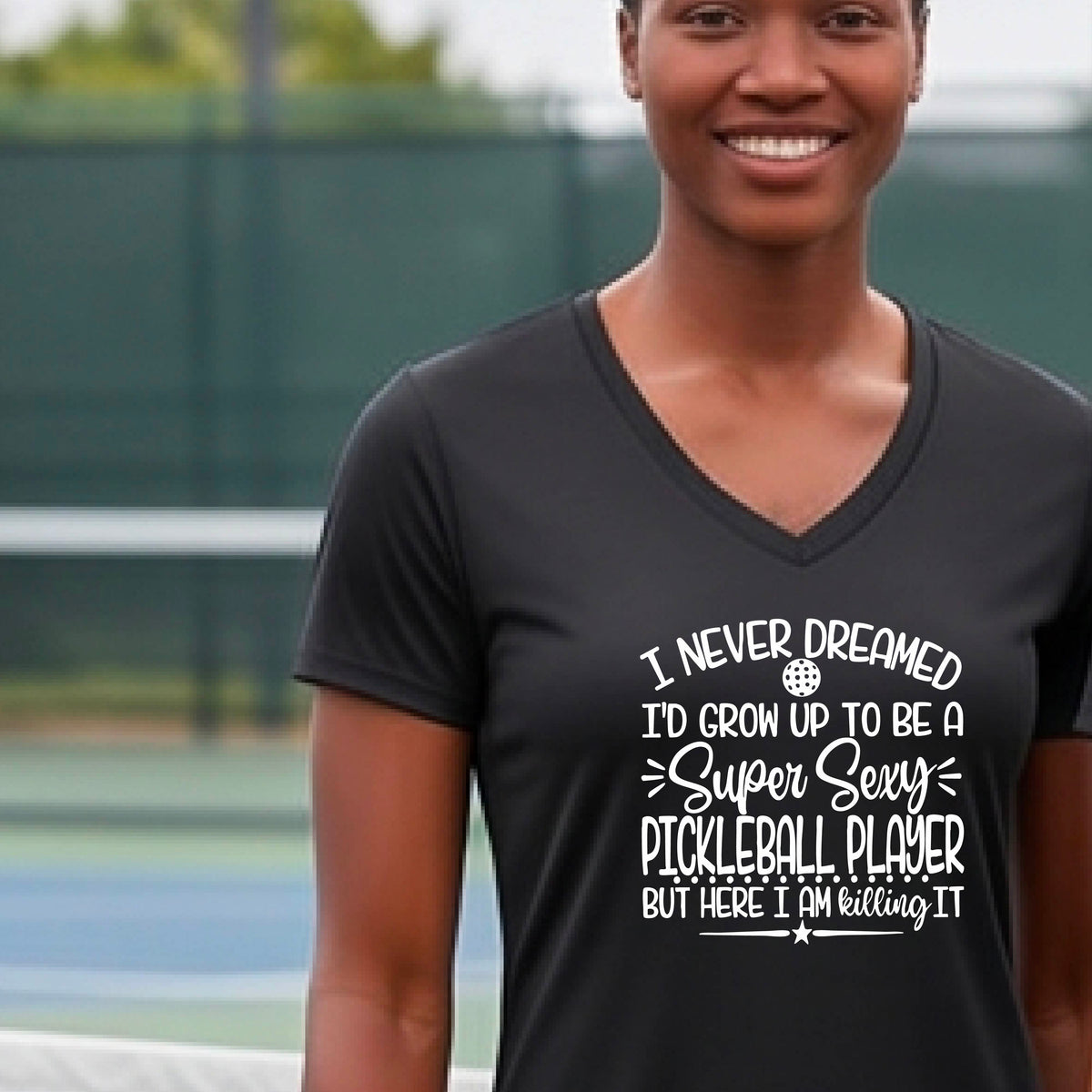 A woman on a pickleball court wearing a black short-sleeve t-shirt with a white graphic of the text:  ‘I Never Dreamed I’d Grow Up To Be A Super Sexy Pickleball Player But Here I Am Killing It!’, standing on a pickleball court.