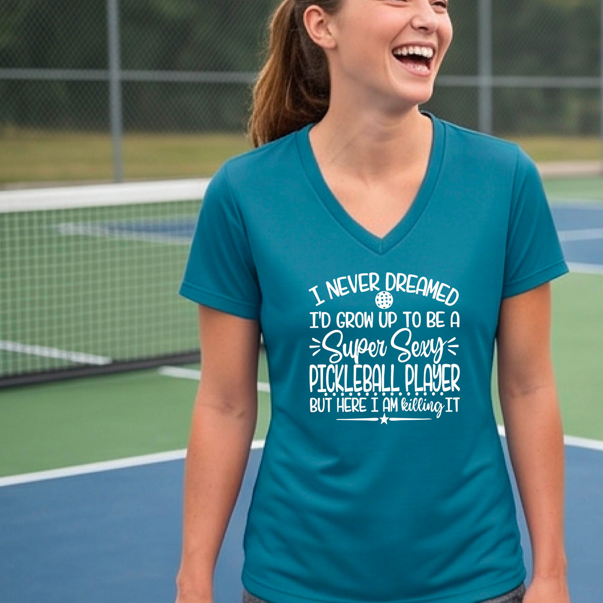 A woman on a pickleball court wearing a teal short-sleeve t-shirt with a white graphic of the text:  ‘I Never Dreamed I’d Grow Up To Be A Super Sexy Pickleball Player But Here I Am Killing It!’, standing on a pickleball court.