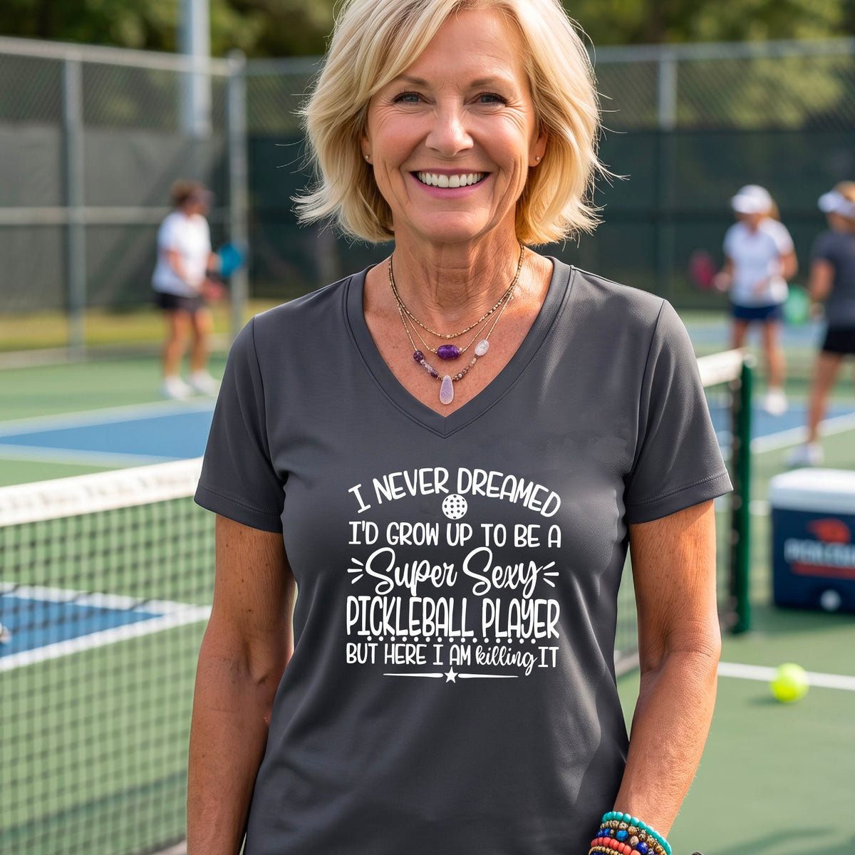 A woman on a pickleball court wearing a gray short-sleeve t-shirt with a white graphic of the text:  ‘I Never Dreamed I’d Grow Up To Be A Super Sexy Pickleball Player But Here I Am Killing It!’, standing on a pickleball court.