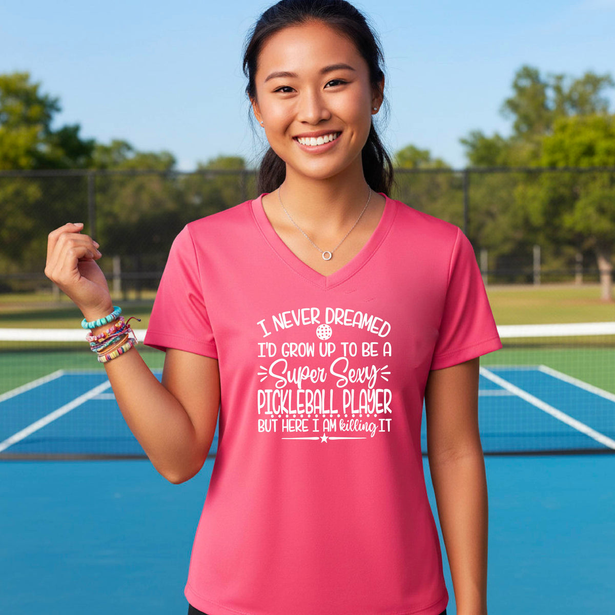 A woman on a pickleball court wearing a coral pink short-sleeve t-shirt with a white graphic of the text:  ‘I Never Dreamed I’d Grow Up To Be A Super Sexy Pickleball Player But Here I Am Killing It!’, standing on a pickleball court.