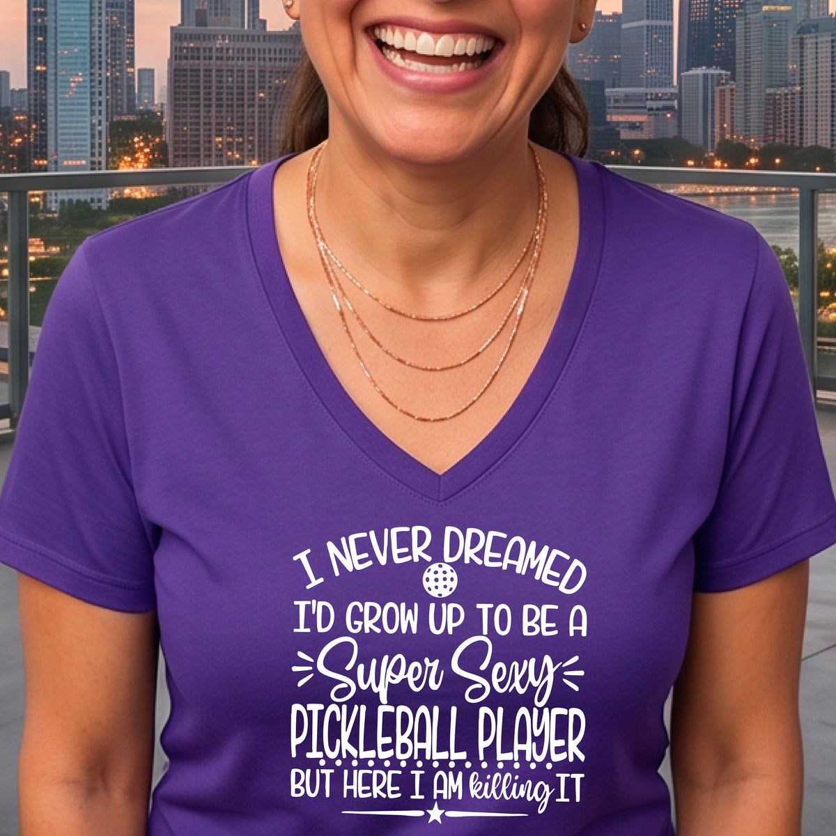 A woman on a pickleball court wearing a purple short-sleeve t-shirt with a white graphic of the text:  ‘I Never Dreamed I’d Grow Up To Be A Super Sexy Pickleball Player But Here I Am Killing It!’, standing on a pickleball court.