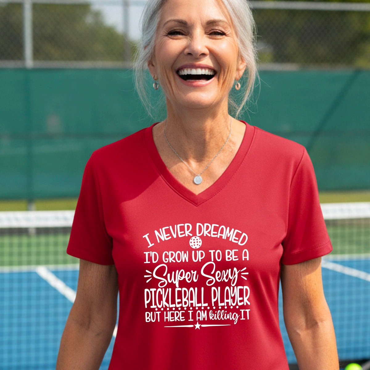 A woman on a pickleball court wearing a red short-sleeve t-shirt with a white graphic of the text:  ‘I Never Dreamed I’d Grow Up To Be A Super Sexy Pickleball Player But Here I Am Killing It!’, standing on a pickleball court.