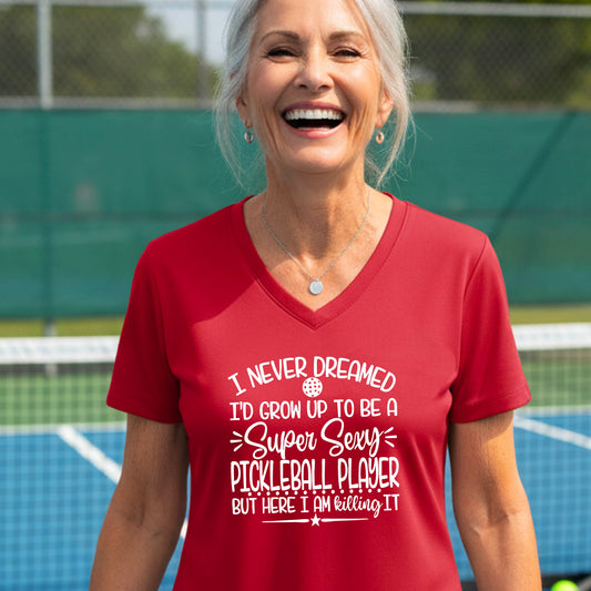 A woman on a pickleball court wearing a red short-sleeve t-shirt with a white graphic of the text:  ‘I Never Dreamed I’d Grow Up To Be A Super Sexy Pickleball Player But Here I Am Killing It!’, standing on a pickleball court.