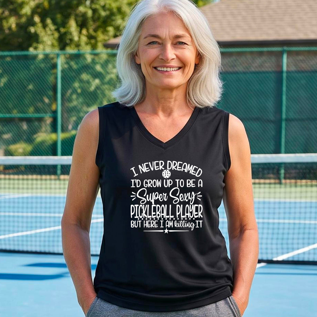A woman on a pickleball court wearing a black sleeveless t-shirt with a white graphic of the text:  ‘I Never Dreamed I’d Grow Up To Be A Super Sexy Pickleball Player But Here I Am Killing It!’, standing on a pickleball court.