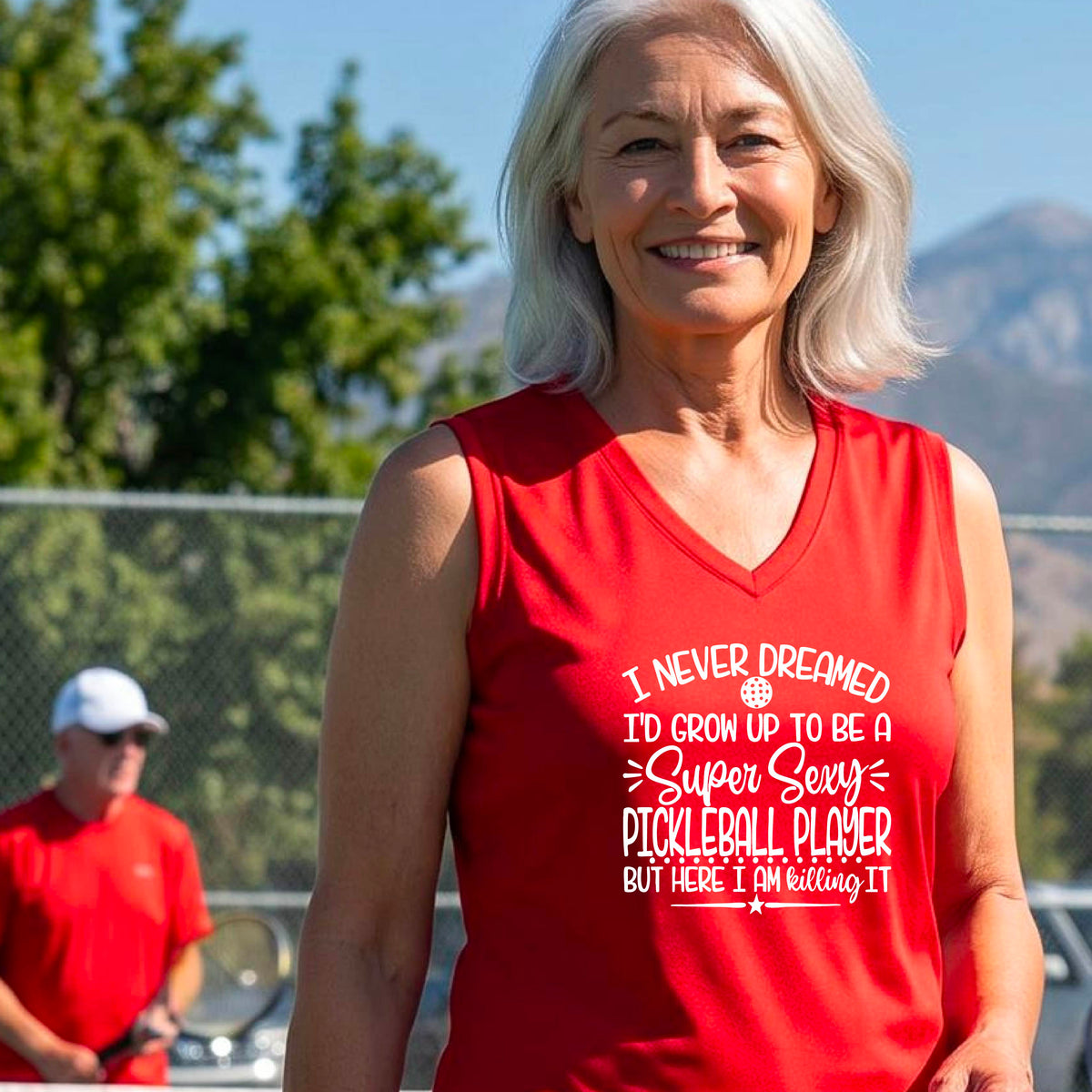 A woman on a pickleball court wearing a red sleeveless t-shirt with a white graphic of the text:  ‘I Never Dreamed I’d Grow Up To Be A Super Sexy Pickleball Player But Here I Am Killing It!’, standing on a pickleball court.