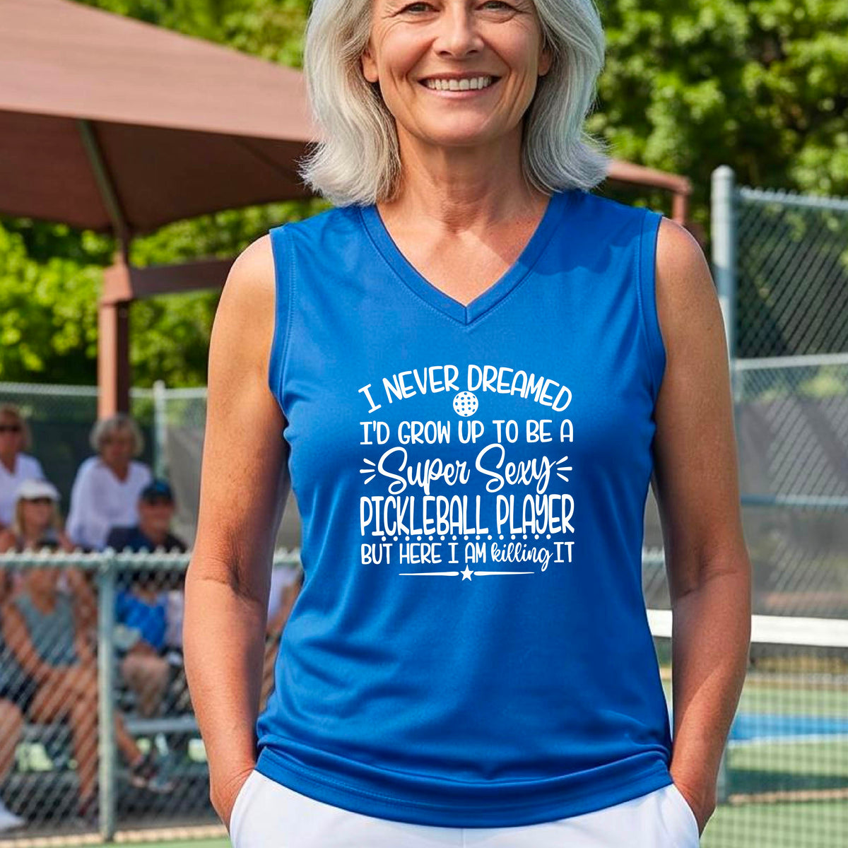 A woman on a pickleball court wearing a royal blue sleeveless t-shirt with a white graphic of the text:  ‘I Never Dreamed I’d Grow Up To Be A Super Sexy Pickleball Player But Here I Am Killing It!’, standing on a pickleball court.