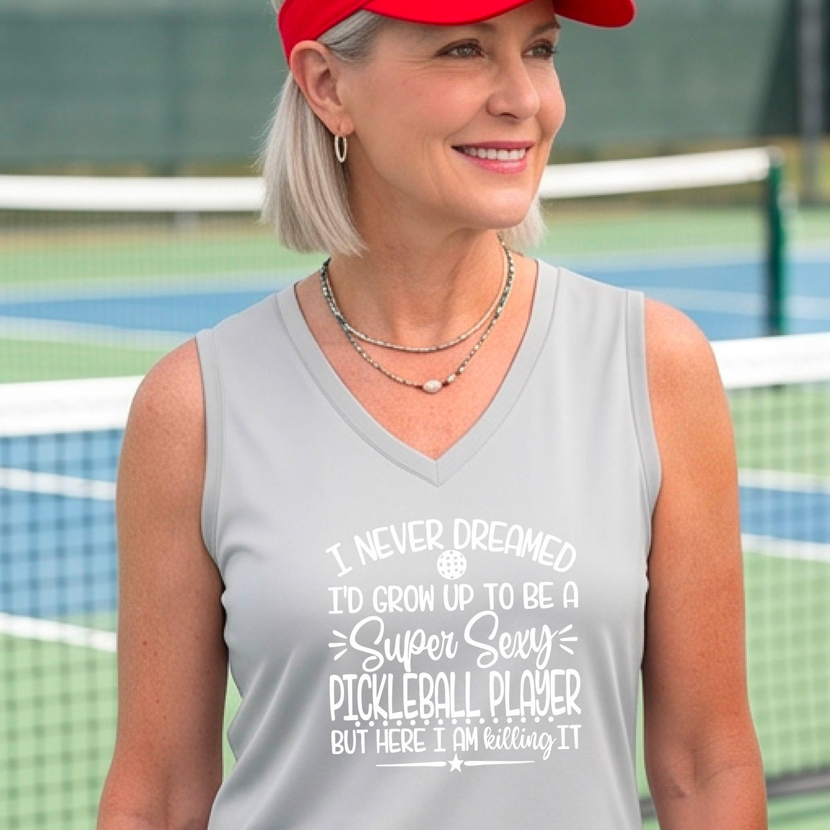 A woman on a pickleball court wearing a silver gray sleeveless t-shirt with a white graphic of the text:  ‘I Never Dreamed I’d Grow Up To Be A Super Sexy Pickleball Player But Here I Am Killing It!’, standing on a pickleball court.