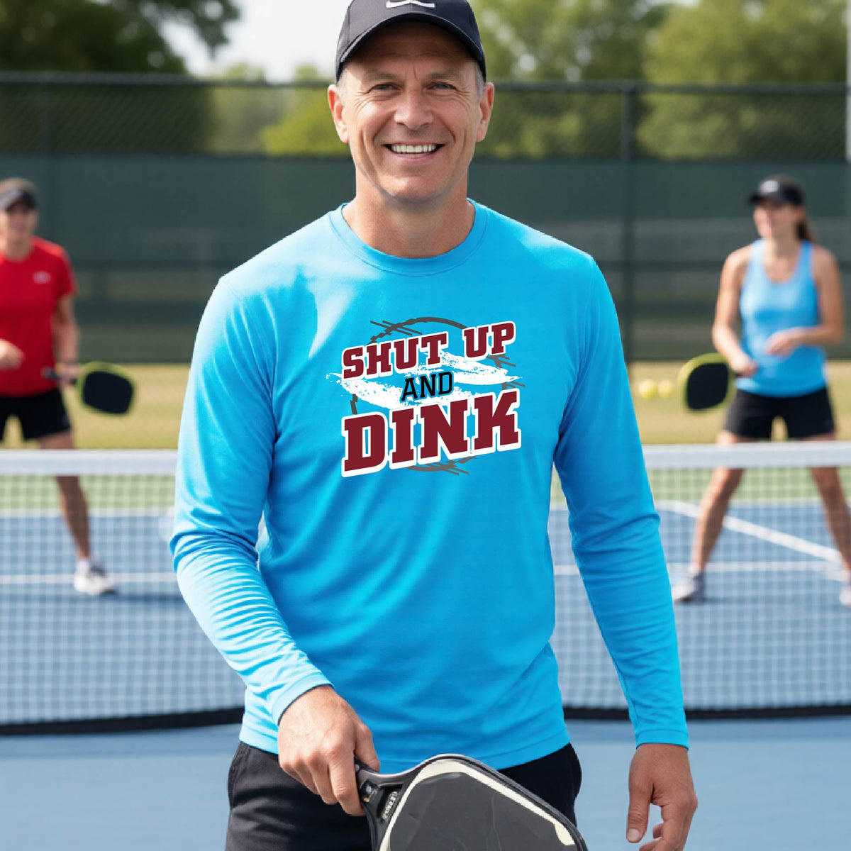 A man on a pickleball court wearing a bright blue long-sleeve t-shirt with a white, gray and black graphic of the text: ‘Shut Up and Dink, standing on a pickleball court.