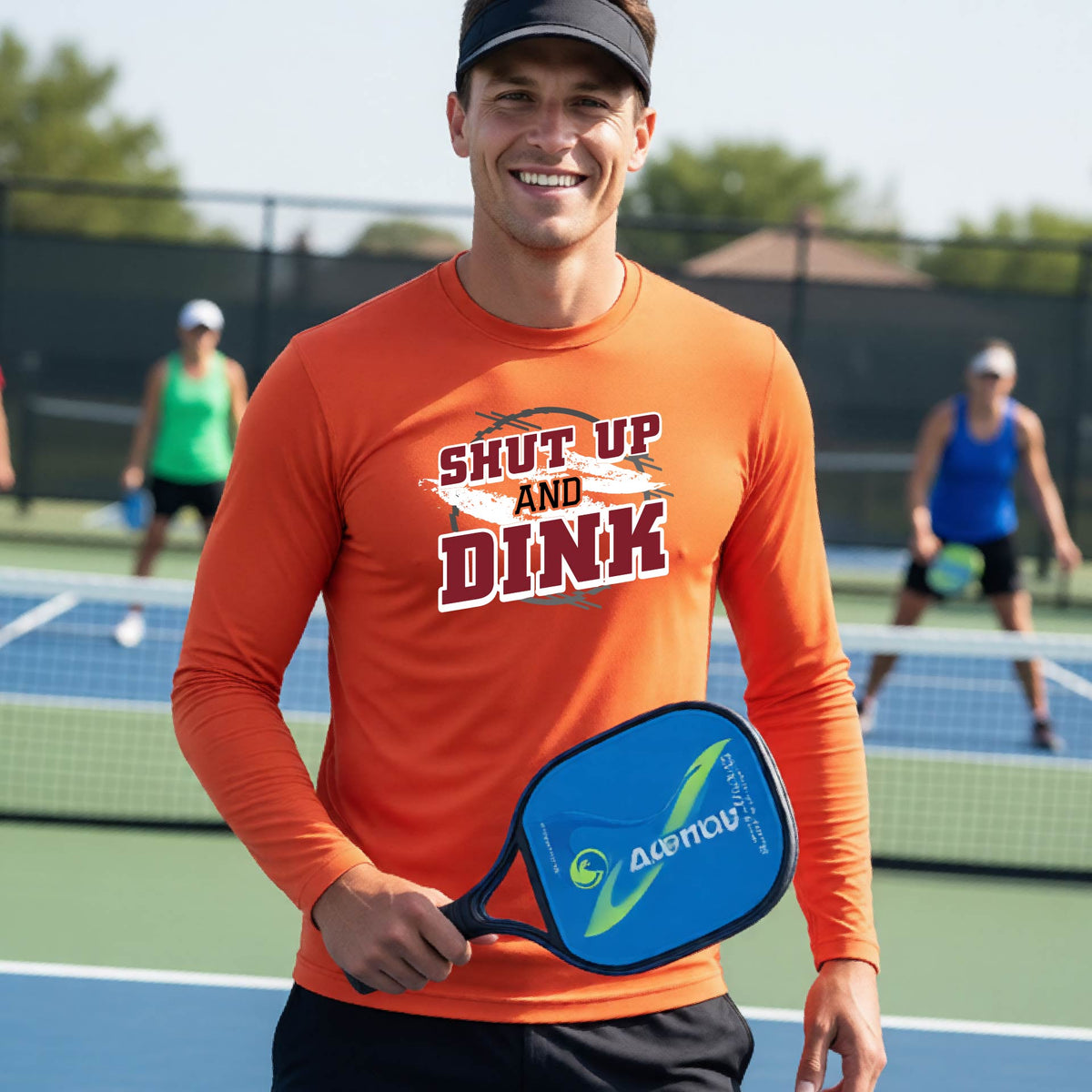 A man on a pickleball court wearing a bright orange long-sleeve t-shirt with a white, gray and black graphic of the text: ‘Shut Up and Dink, standing on a pickleball court.