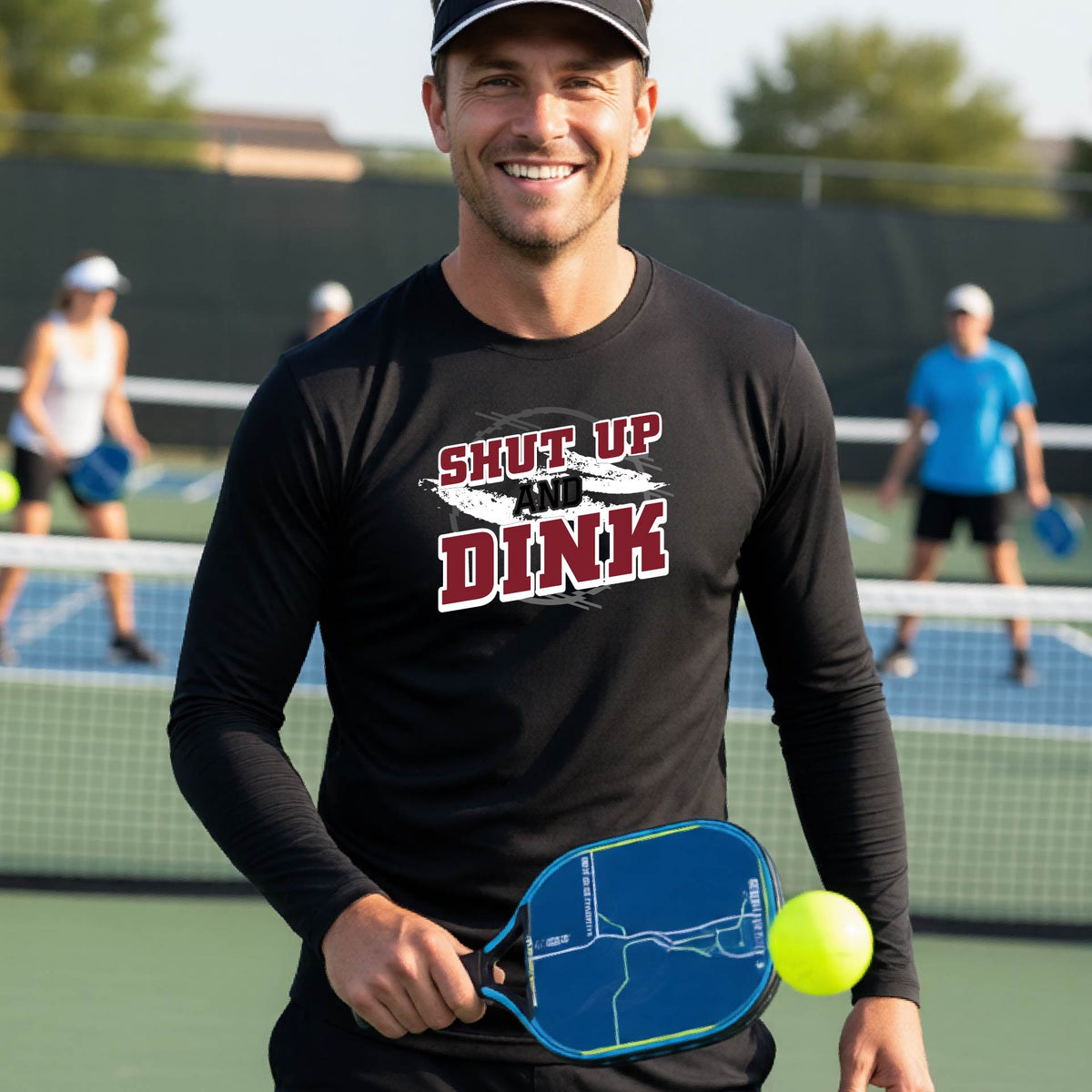 A man on a pickleball court wearing a black long-sleeve t-shirt with a white, gray and black graphic of the text: ‘Shut Up and Dink, standing on a pickleball court.
