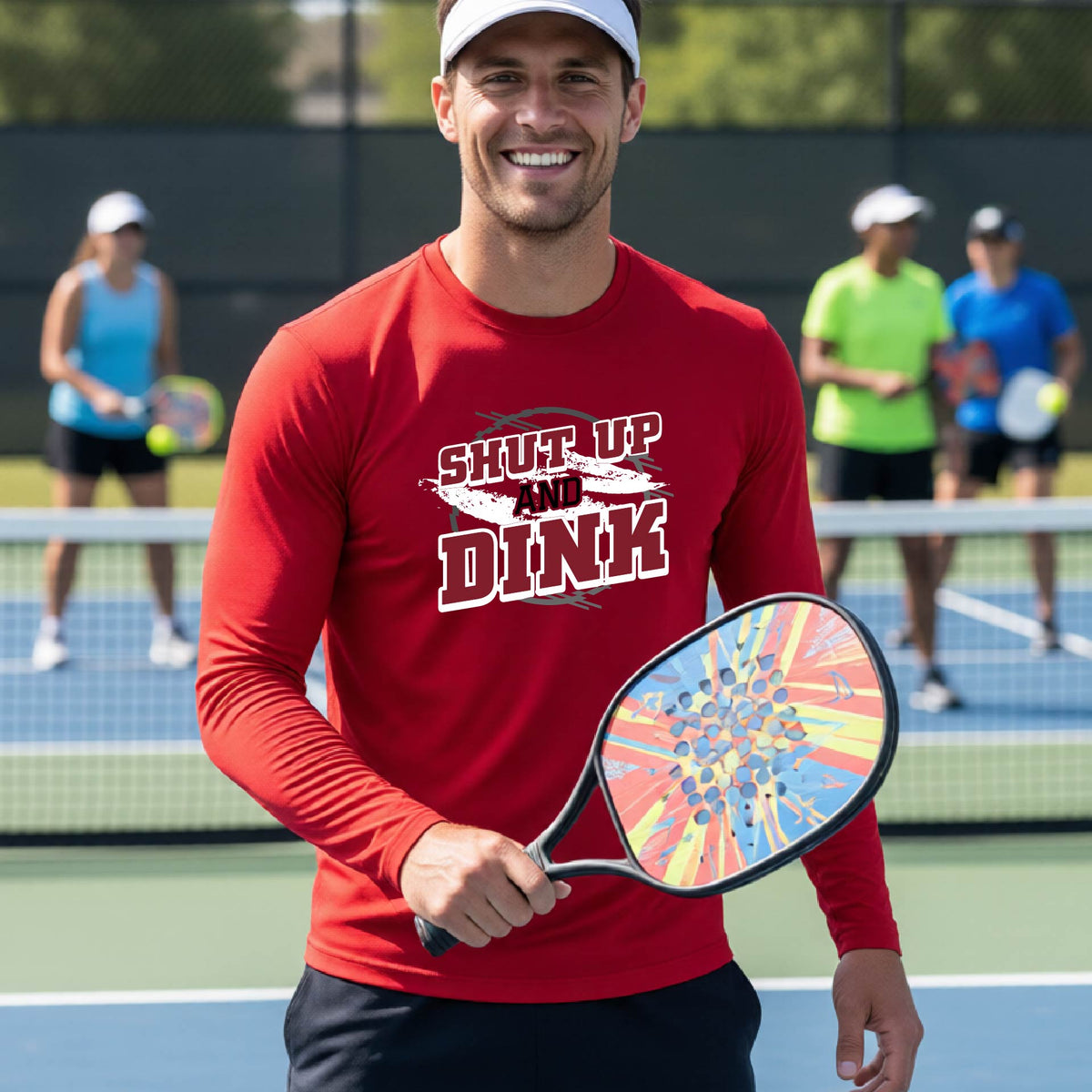 A man on a pickleball court wearing a red long-sleeve t-shirt with a white, gray and black graphic of the text: ‘Shut Up and Dink, standing on a pickleball court.