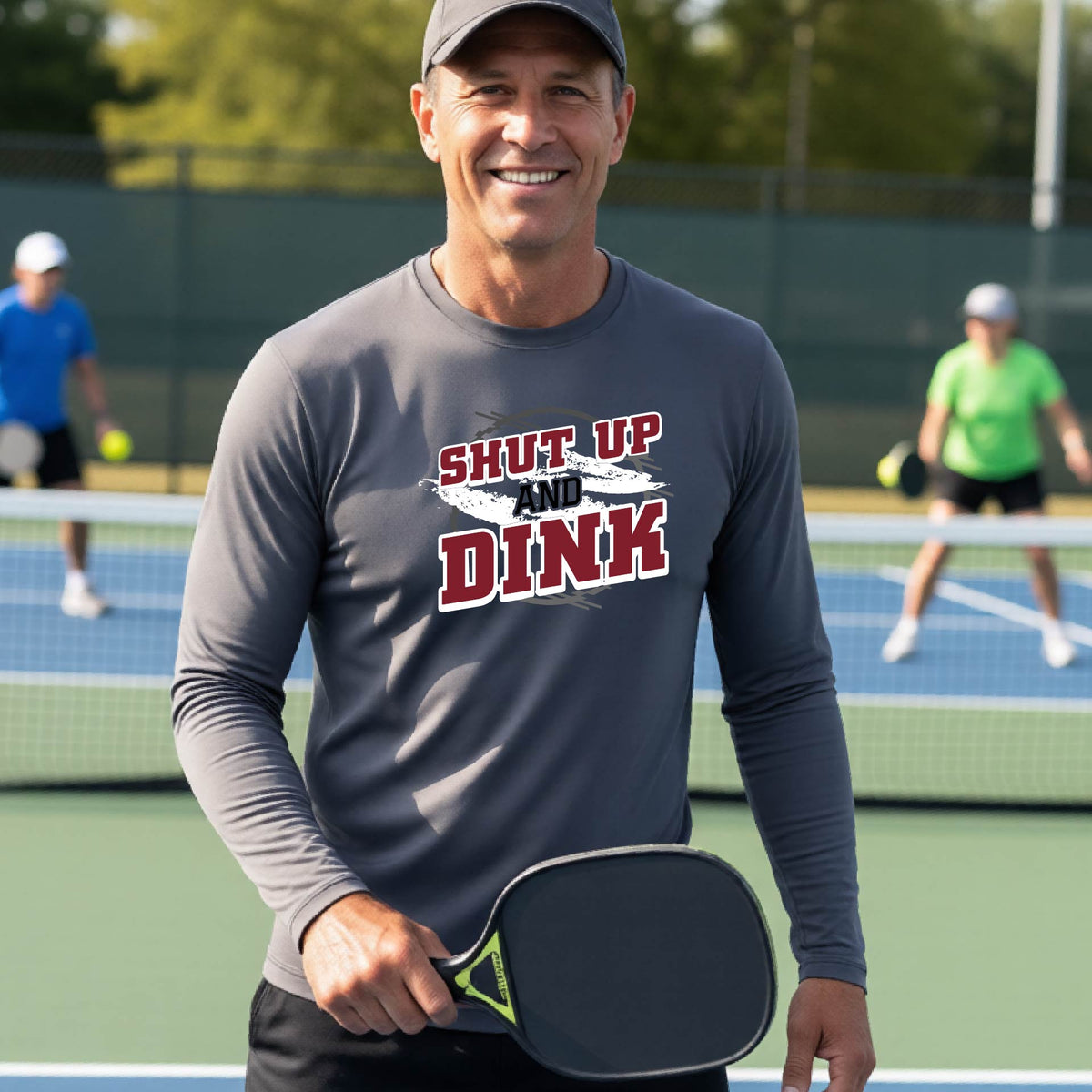 dark g long-sleeve t-shirt with a white, dark gray and black graphic of the text: ‘Shut Up and Dink, standing on a pickleball court.