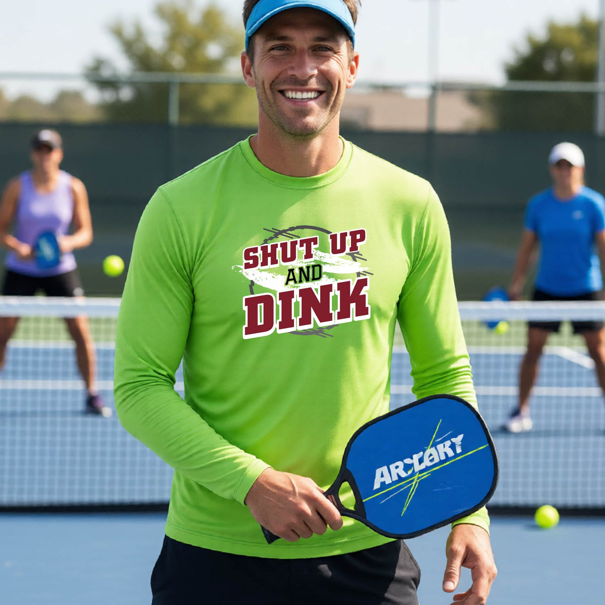 A man on a pickleball court wearing a lime green long-sleeve t-shirt with a white, gray and black graphic of the text: ‘Shut Up and Dink, standing on a pickleball court.