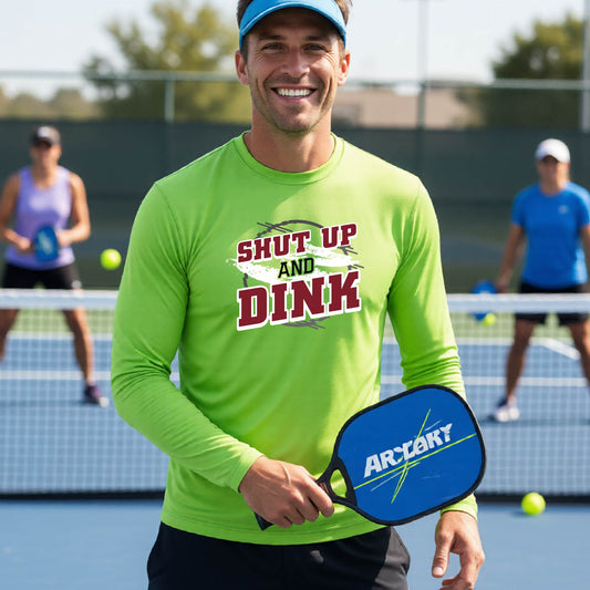 A man on a pickleball court wearing a lime green long-sleeve t-shirt with a white, gray and black graphic of the text: ‘Shut Up and Dink, standing on a pickleball court.