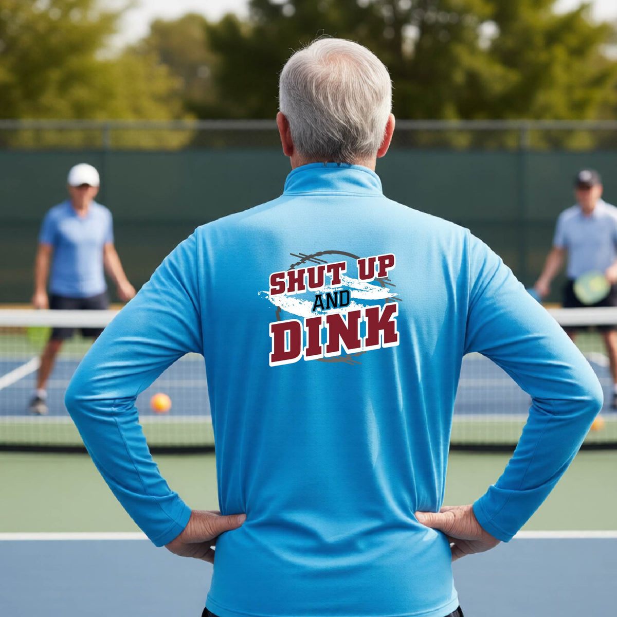 A man on a pickleball court, facing backwards, wearing a bright blue quarter-zip shirt with a white, gray and black graphic of the text: ‘Shut Up and Dink, standing on a pickleball court.