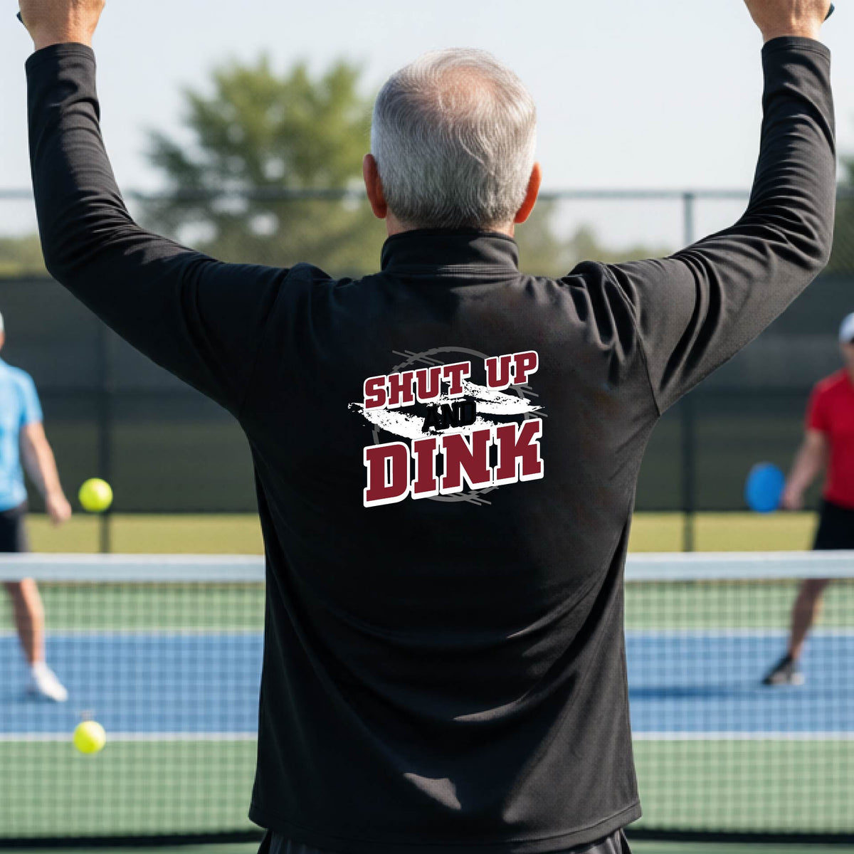 A man on a pickleball court, facing backwards, wearing a black quarter-zip shirt with a white, gray and black graphic of the text: ‘Shut Up and Dink, standing on a pickleball court.
