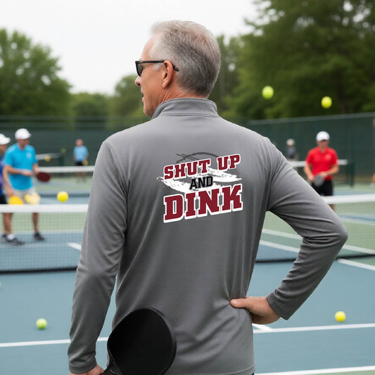 A man on a pickleball court, facing backwards, wearing a gray quarter-zip shirt with a white, gray and black graphic of the text: ‘Shut Up and Dink, standing on a pickleball court.