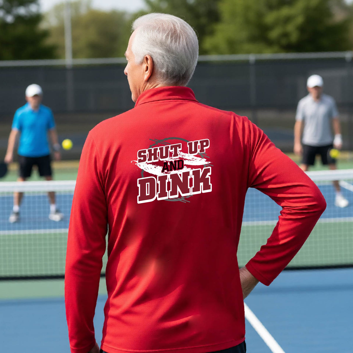 A man on a pickleball court, facing backwards, wearing a red quarter-zip shirt with a white, gray and black graphic of the text: ‘Shut Up and Dink, standing on a pickleball court.