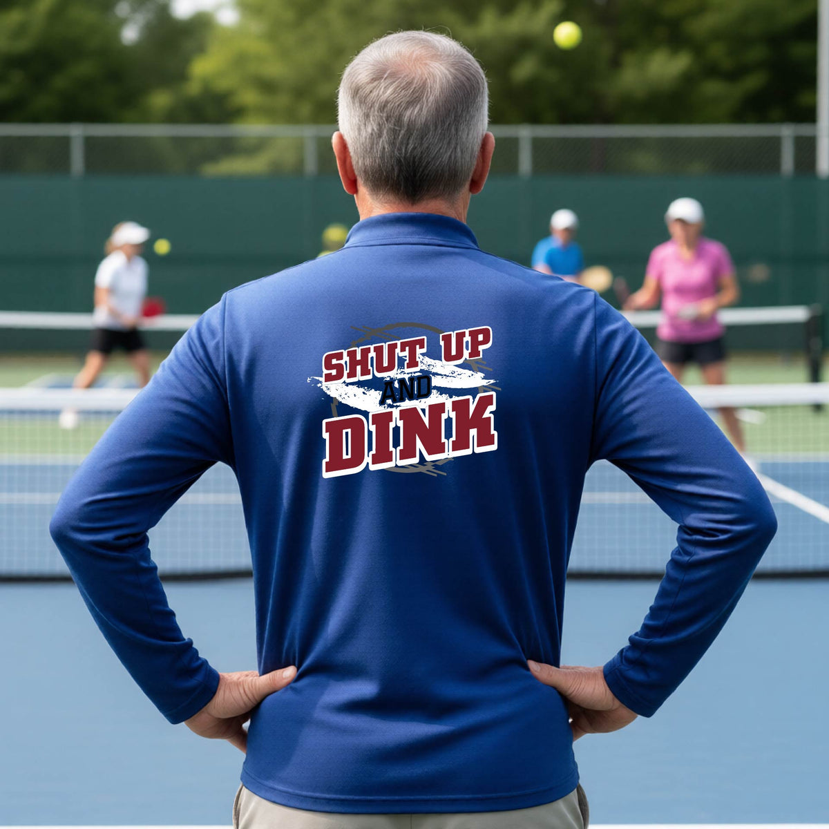 A man on a pickleball court, facing backwards, wearing a royal blue quarter-zip shirt with a white, gray and black graphic of the text: ‘Shut Up and Dink, standing on a pickleball court.