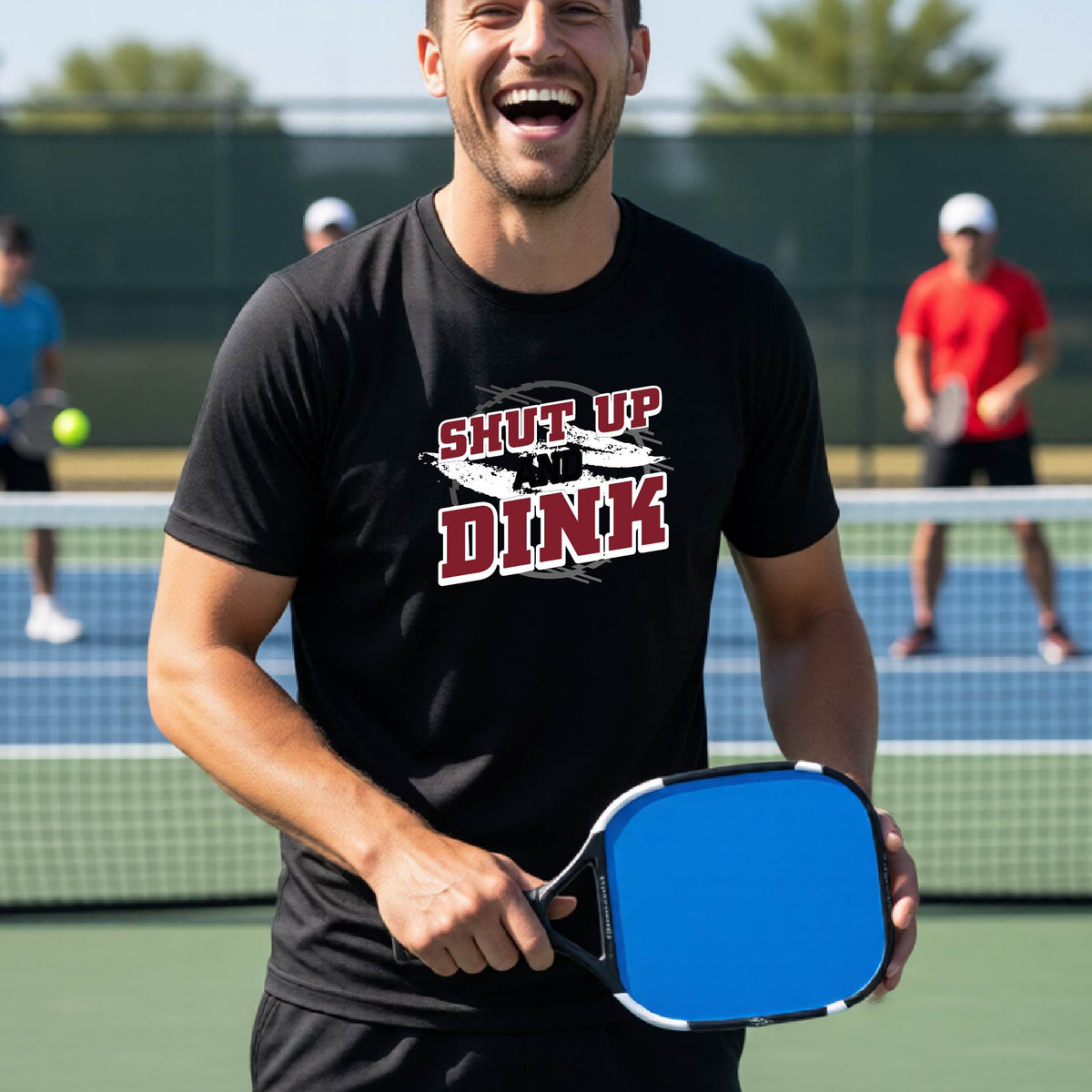 A man on a pickleball court wearing a black long-sleeve t-shirt with a white, gray and black graphic of the text: ‘Shut Up and Dink, standing on a pickleball court.