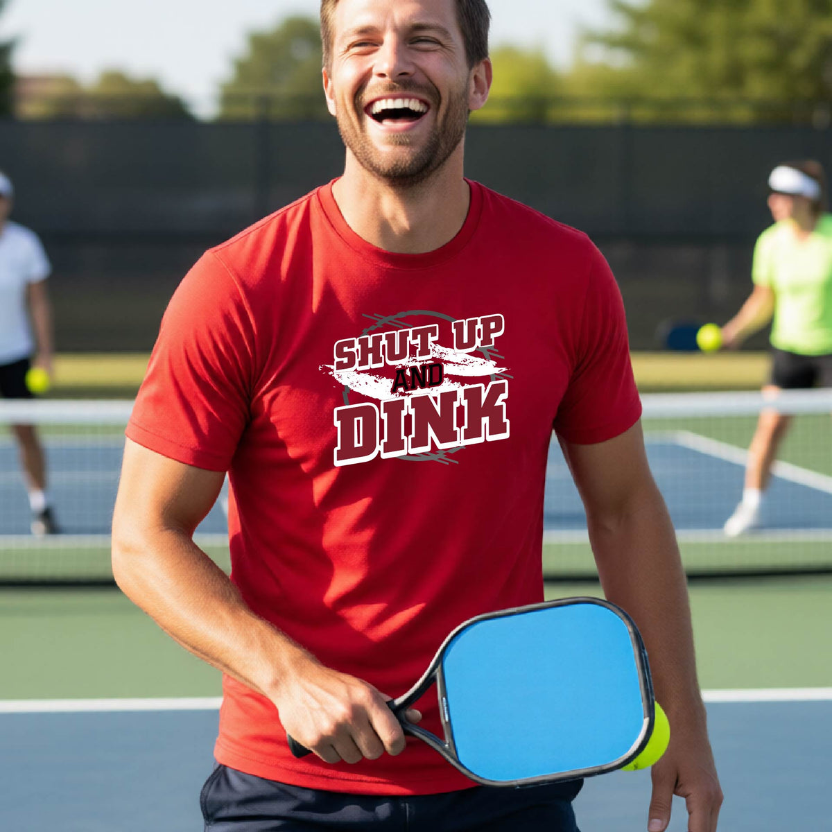 A man on a pickleball court wearing a red long-sleeve t-shirt with a white, gray and black graphic of the text: ‘Shut Up and Dink, standing on a pickleball court.
