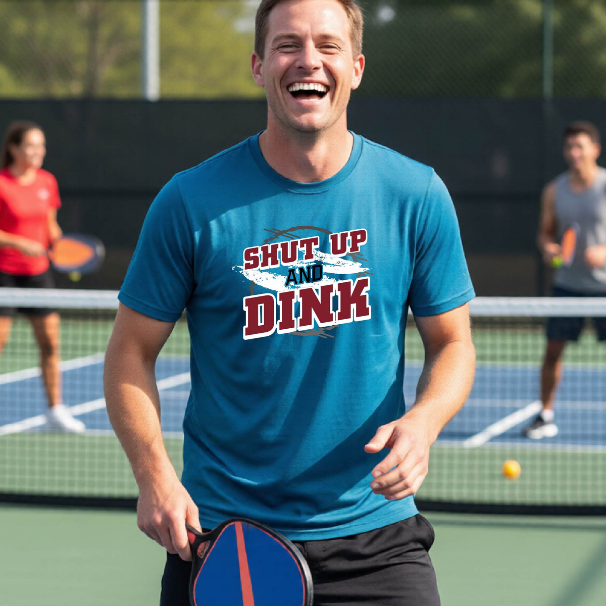 A man on a pickleball court wearing a teal long-sleeve t-shirt with a white, gray and black graphic of the text: ‘Shut Up and Dink, standing on a pickleball court.