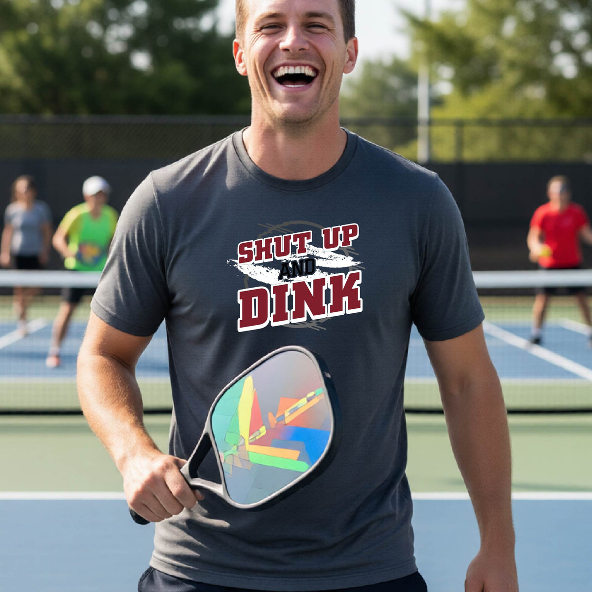 A man on a pickleball court wearing a dark gray long-sleeve t-shirt with a white, gray and black graphic of the text: ‘Shut Up and Dink, standing on a pickleball court.