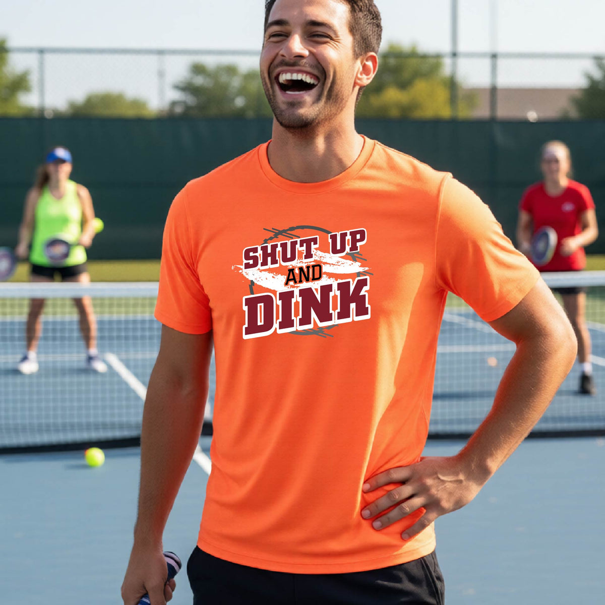A man on a pickleball court wearing a bright orange long-sleeve t-shirt with a white, gray and black graphic of the text: ‘Shut Up and Dink, standing on a pickleball court.