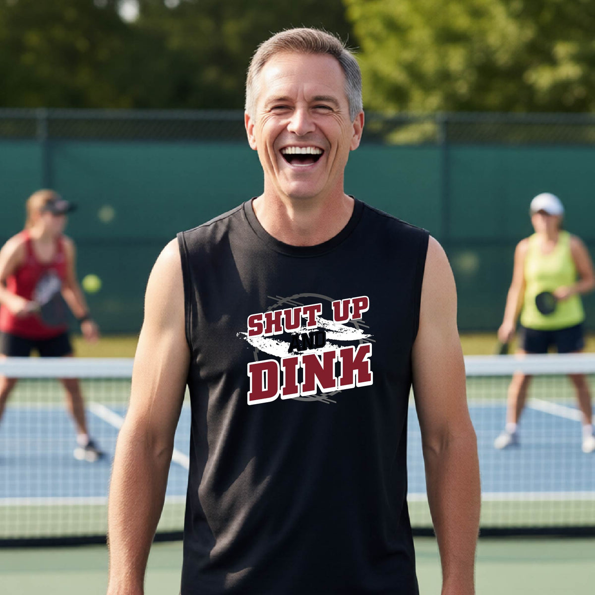 A man on a pickleball court wearing a black sleeveless t-shirt with a white, gray and black graphic of the text: ‘Shut Up and Dink, standing on a pickleball court.