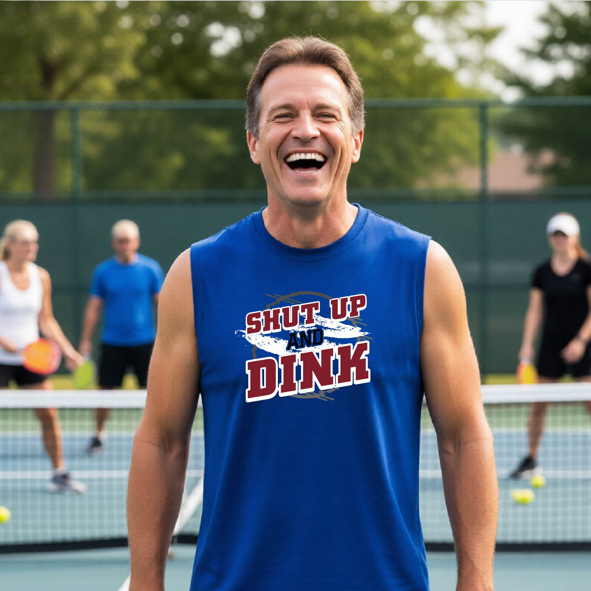 A man on a pickleball court wearing a royal blue sleeveless t-shirt with a white, gray and black graphic of the text: ‘Shut Up and Dink, standing on a pickleball court.