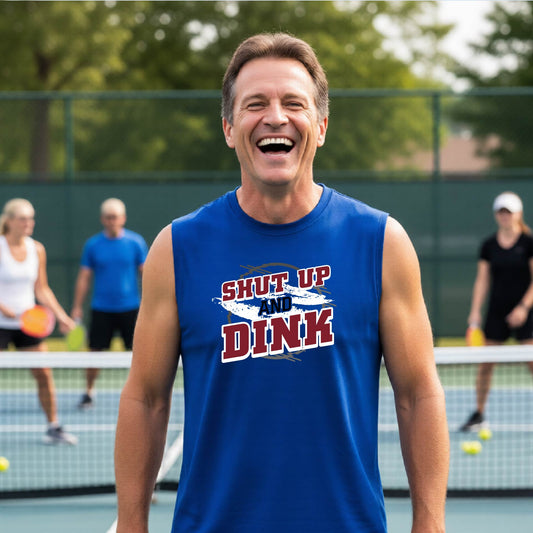 A man on a pickleball court wearing a royal blue sleeveless t-shirt with a white, gray and black graphic of the text: ‘Shut Up and Dink, standing on a pickleball court.