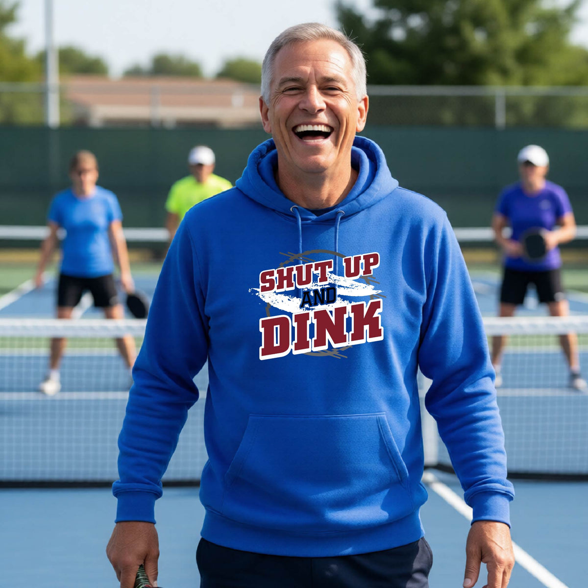 A man on a pickleball court wearing a royal blue unisex pullover hoodie with a white, gray and black graphic of the text: ‘Shut Up and Dink, standing on a pickleball court.