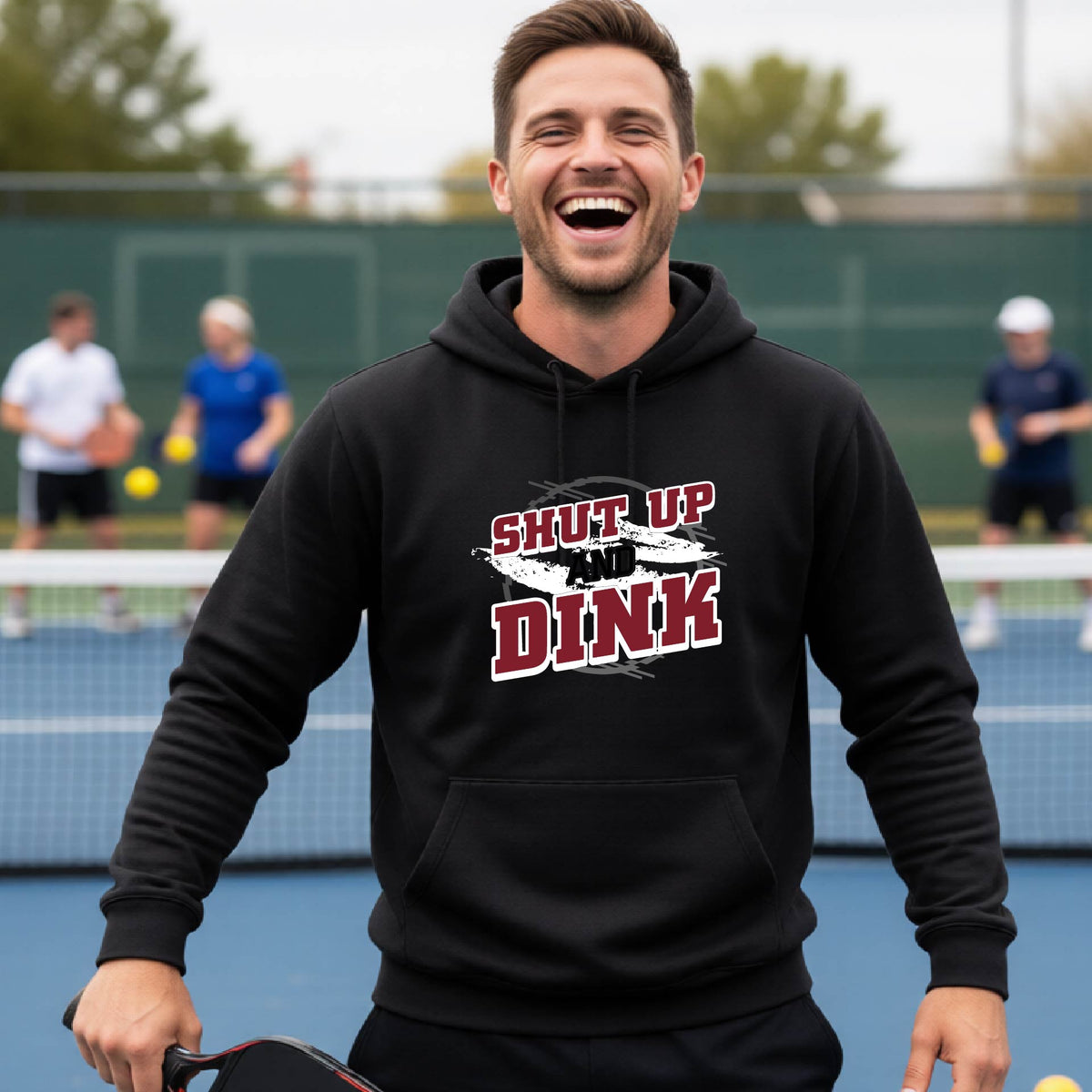A man on a pickleball court wearing a black unisex pullover hoodie with a white, gray and black graphic of the text: ‘Shut Up and Dink, standing on a pickleball court.