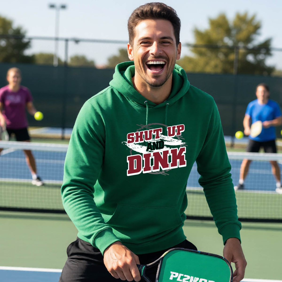 A man on a pickleball court wearing a green unisex pullover hoodie with a white, gray and black graphic of the text: ‘Shut Up and Dink, standing on a pickleball court.