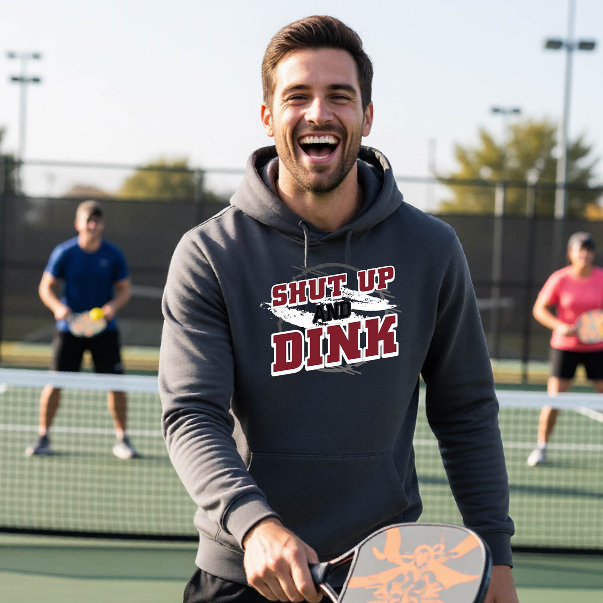 A man on a pickleball court wearing a dark gray unisex pullover hoodie with a white, gray and black graphic of the text: ‘Shut Up and Dink, standing on a pickleball court.