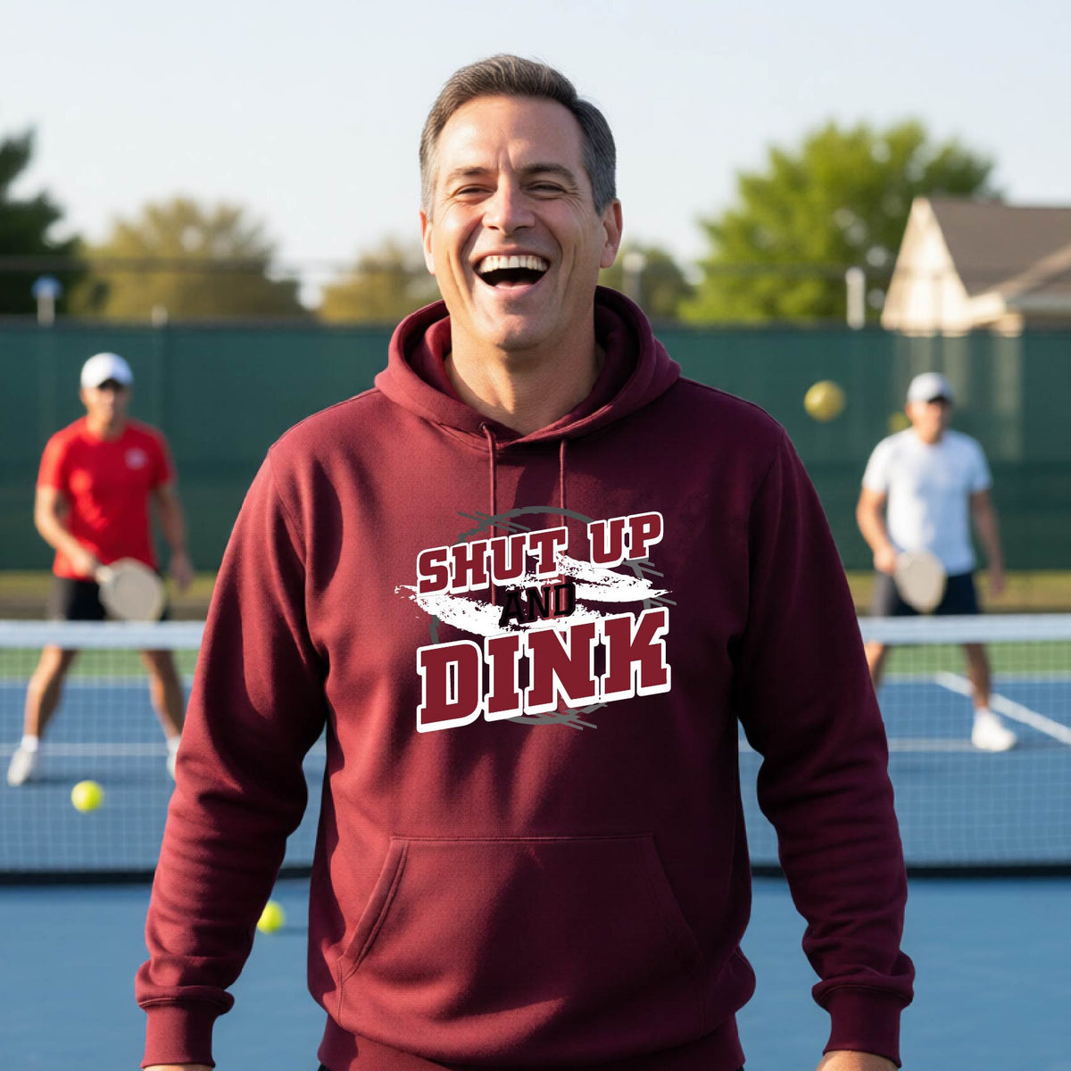A man on a pickleball court wearing a black unisex pullover hoodie with a white, gray and black graphic of the text: ‘Shut Up and Dink, standing on a pickleball court.