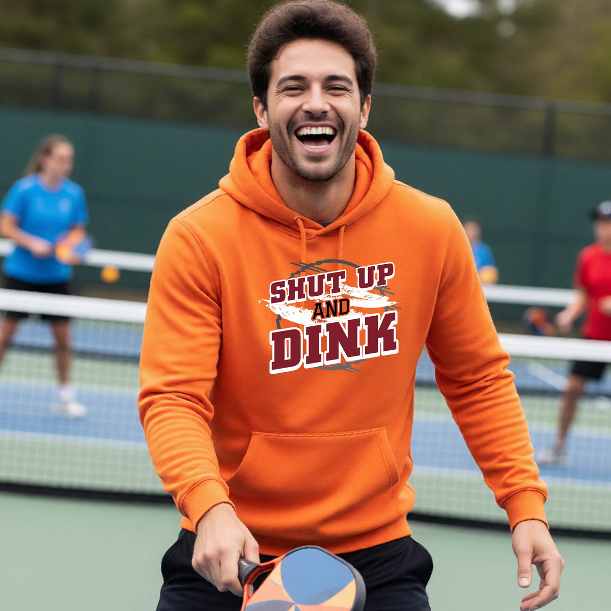 A man on a pickleball court wearing a bright orange unisex pullover hoodie with a white, gray and black graphic of the text: ‘Shut Up and Dink, standing on a pickleball court.