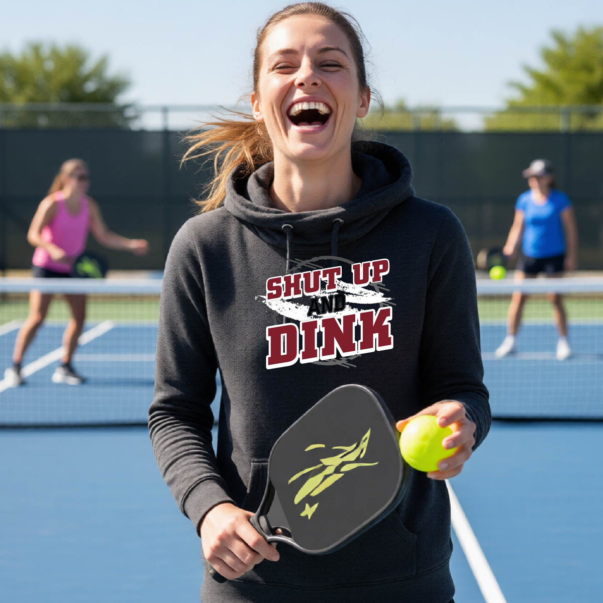 A woman on a pickleball court wearing a black cowl-neck pullover hoodie with a white, gray and black graphic of the text: ‘Shut Up and Dink, standing on a pickleball court.