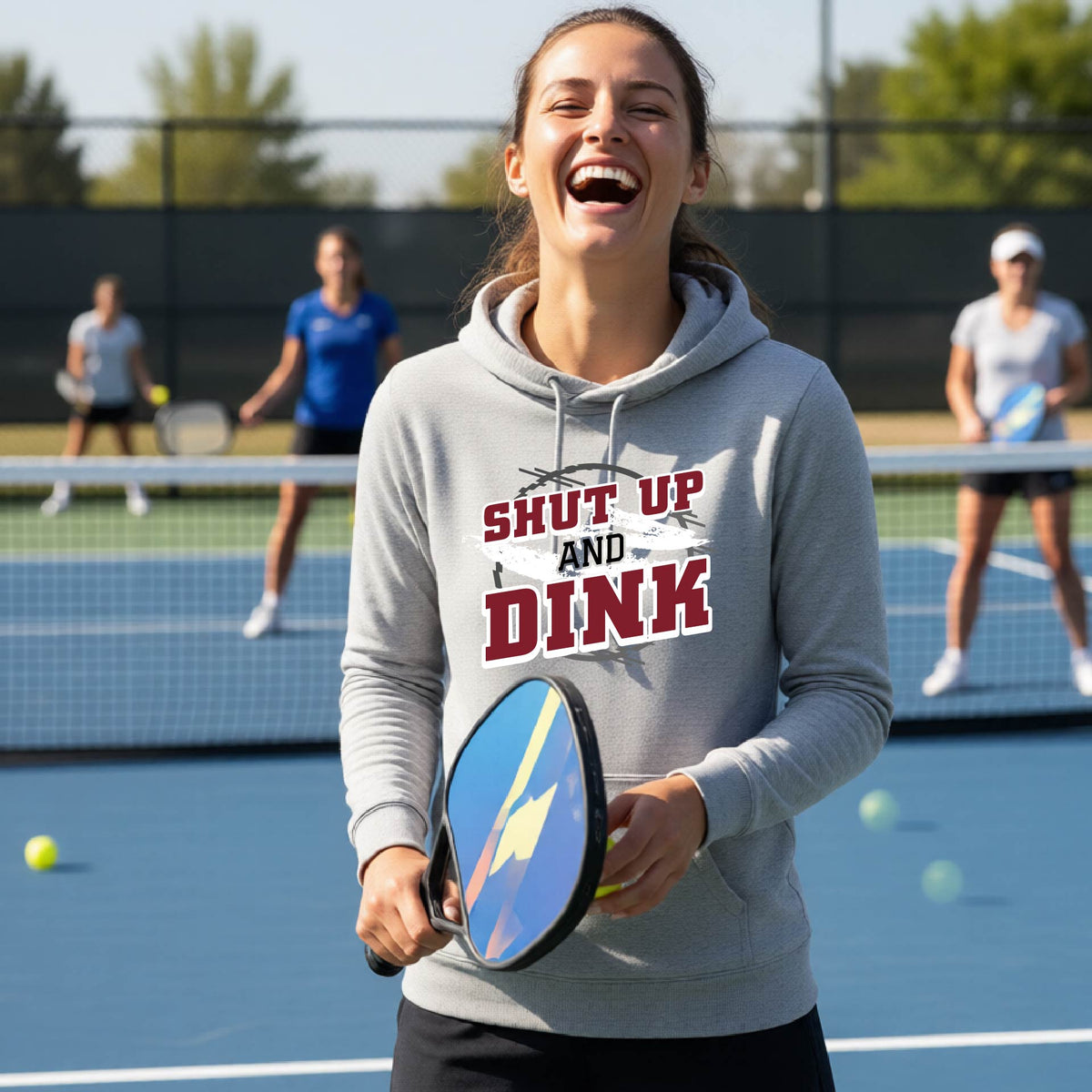 A woman on a pickleball court wearing a gray cowl-neck pullover hoodie with a white, gray and black graphic of the text: ‘Shut Up and Dink, standing on a pickleball court.