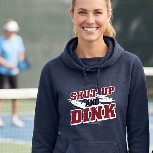 A woman on a pickleball court wearing a navy blue cowl-neck pullover hoodie with a white, gray and black graphic of the text: ‘Shut Up and Dink, standing on a pickleball court.