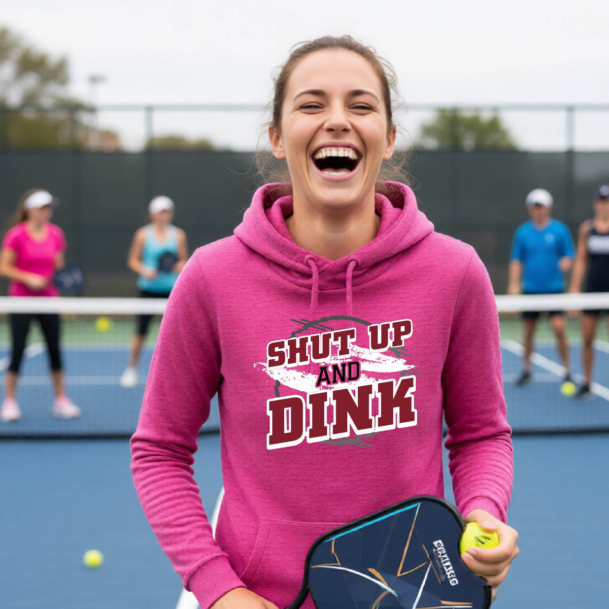 A woman on a pickleball court wearing a pink cowl-neck pullover hoodie with a white, gray and black graphic of the text: ‘Shut Up and Dink, standing on a pickleball court.