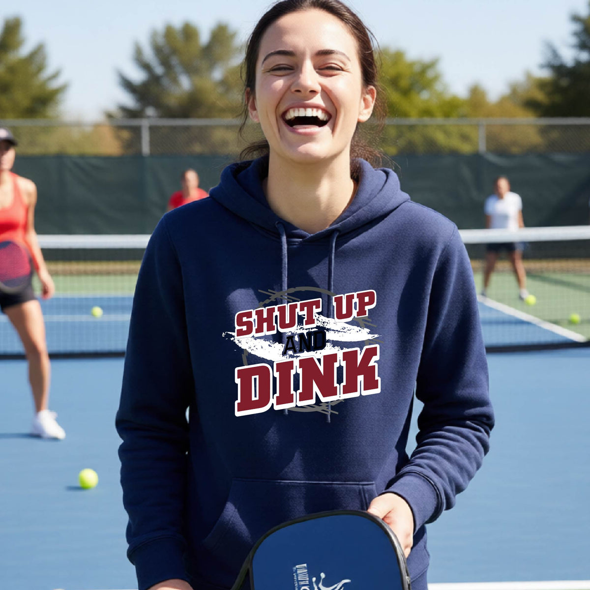 A woman on a pickleball court wearing a navy blue hoodie pullover with a white, gray and black graphic of the text: ‘Shut Up and Dink, standing on a pickleball court.