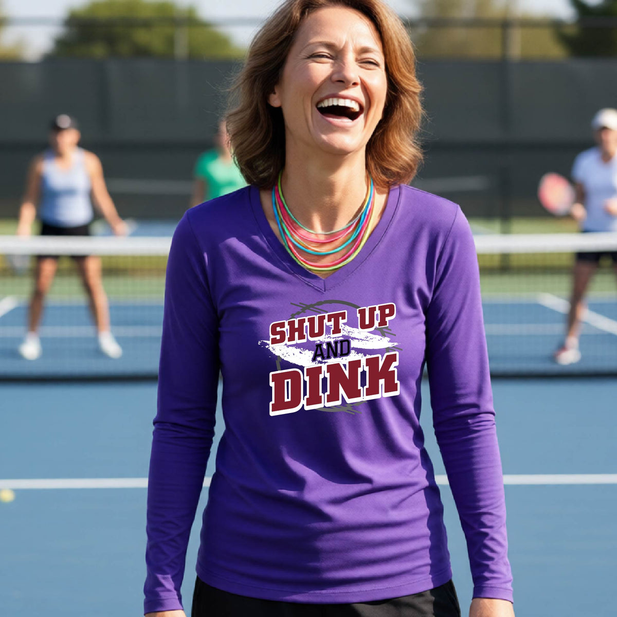 A woman on a pickleball court wearing a purple long-sleeve t-shirt with a white, gray and black graphic of the text: ‘Shut Up and Dink, standing on a pickleball court.
