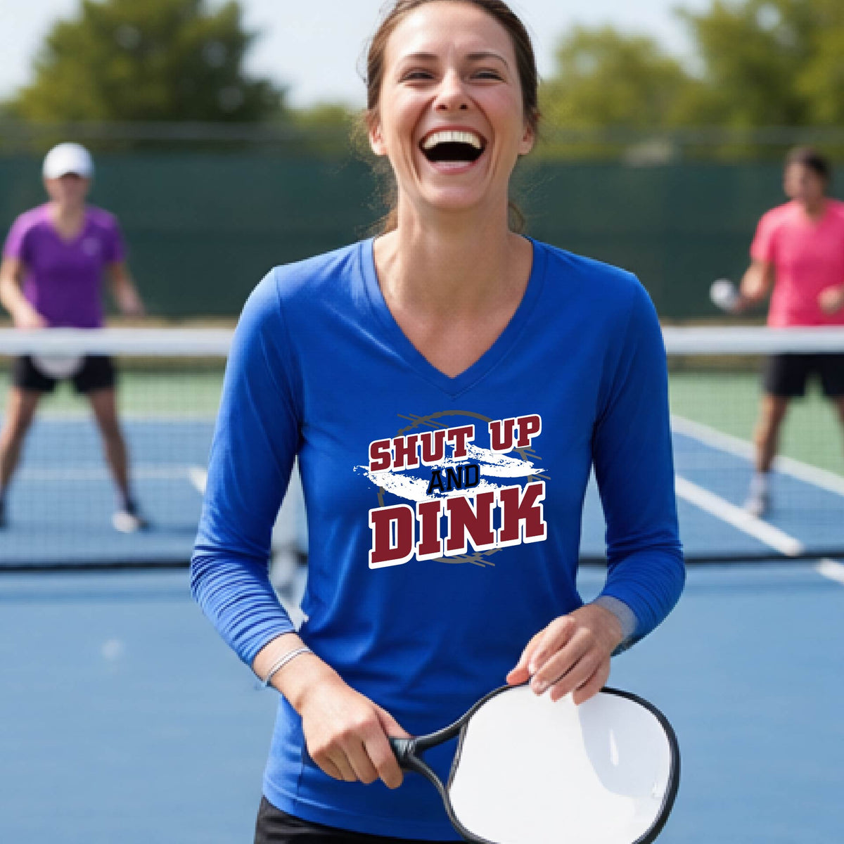 A woman on a pickleball court wearing a royal blue long-sleeve t-shirt with a white, gray and black graphic of the text: ‘Shut Up and Dink, standing on a pickleball court.