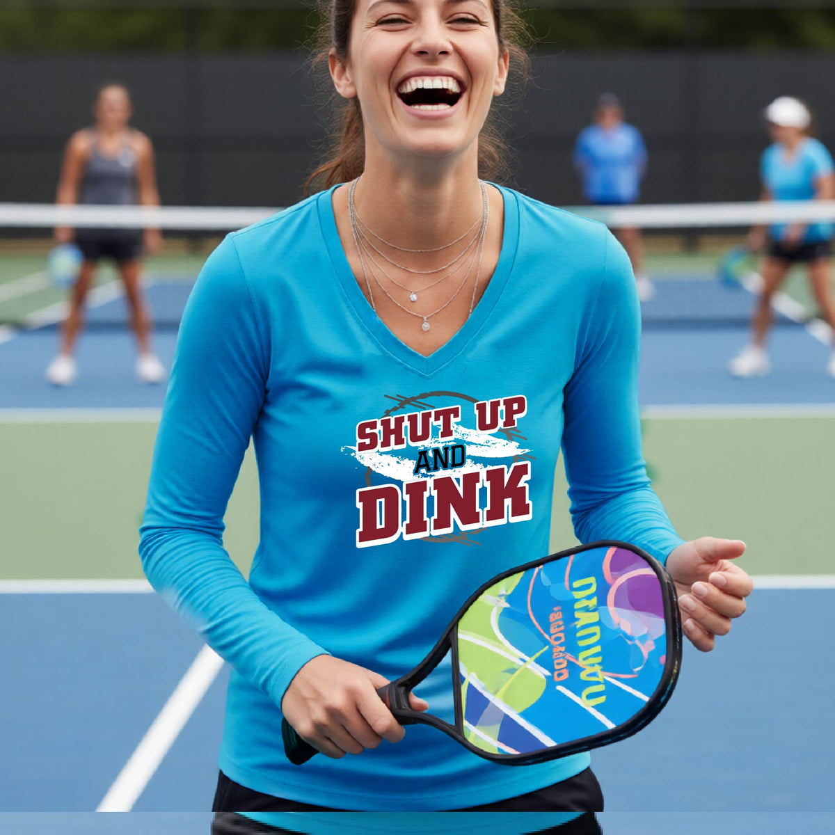 A woman on a pickleball court wearing a bright blue long-sleeve t-shirt with a white, gray and black graphic of the text: ‘Shut Up and Dink, standing on a pickleball court.