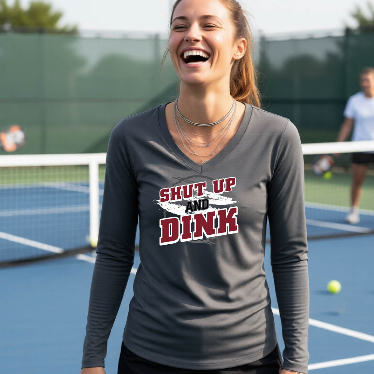 A woman on a pickleball court wearing a dark gray long-sleeve t-shirt with a white, gray and black graphic of the text: ‘Shut Up and Dink, standing on a pickleball court.